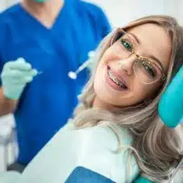 Smiling female patient wearing glasses in dental chair with dentist in blue scrubs behind her.