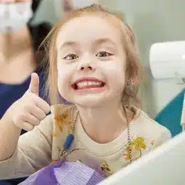 Young girl with red hair smiling at dental office, giving thumbs up gesture.