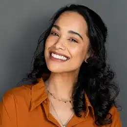 Smiling woman with long black hair wearing orange shirt against gray background, dental professional portrait.
